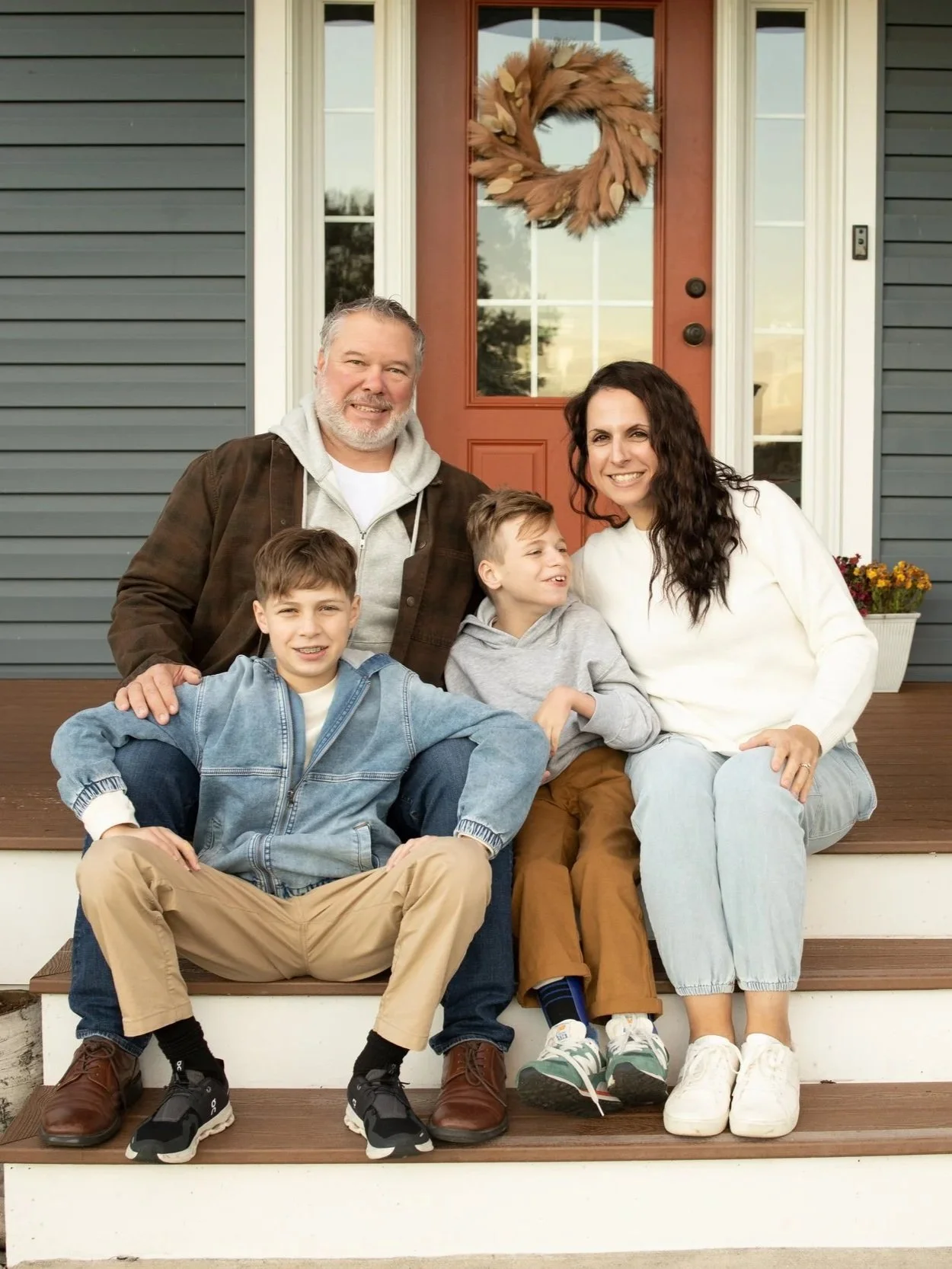 A family of four sitting on the front porch of their house, smiling at the camera. The family includes an older man, a woman, and two young boys. The house has gray siding, a red door with a wreath, and a flower box with yellow flowers.