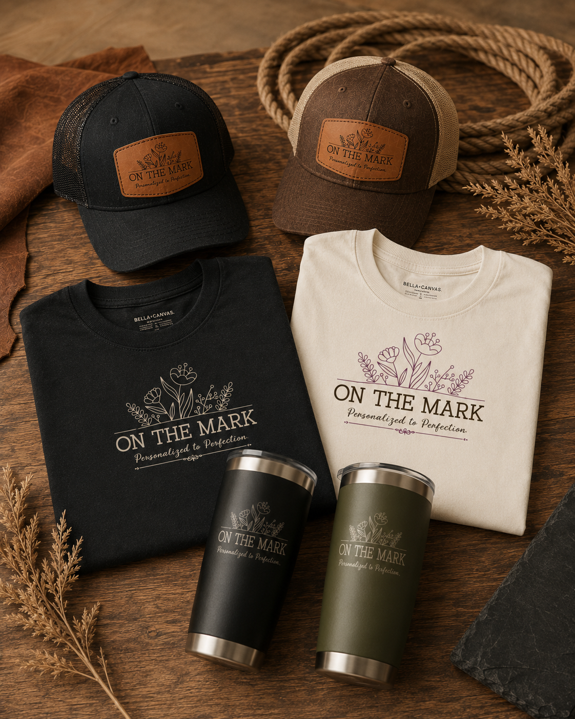 Two baseball caps, one black and one brown, with leather patches reading 'On The Mark.' Two folded T-shirts, one black and one cream, both with a floral design and the phrase 'On The Mark Personalized to Perfection.' Two reusable tumblers, one black and one green, with matching floral and text designs. Items are arranged on a wooden surface with a coiled rope, dried leaves, and a stone nearby.