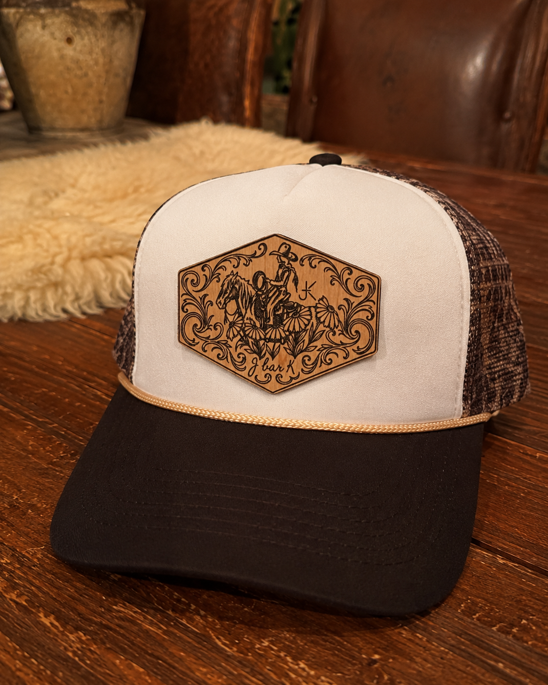 A trucker cap with a white front panel, black brim, and a decorative patch featuring a cowboy on a horse, floral designs, and the text 'J bar K'.