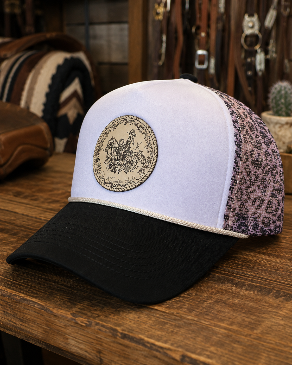 A white and black trucker hat with a decorative patch on the front, sitting on a wooden table in a rustic store with leather belts and a cactus in the background.