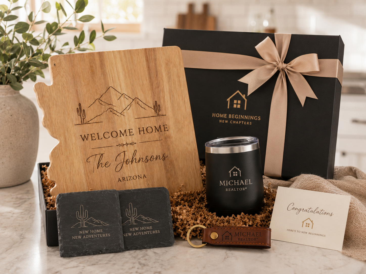 A welcome home gift set including a wooden plaque with mountains, cacti, and the word 'Welcome Home,' a black gift box with a beige ribbon, personalized tumblers with a house logo, a leather keychain, guest towels, a potted plant, and a congratulatory card, all on a light-colored countertop.