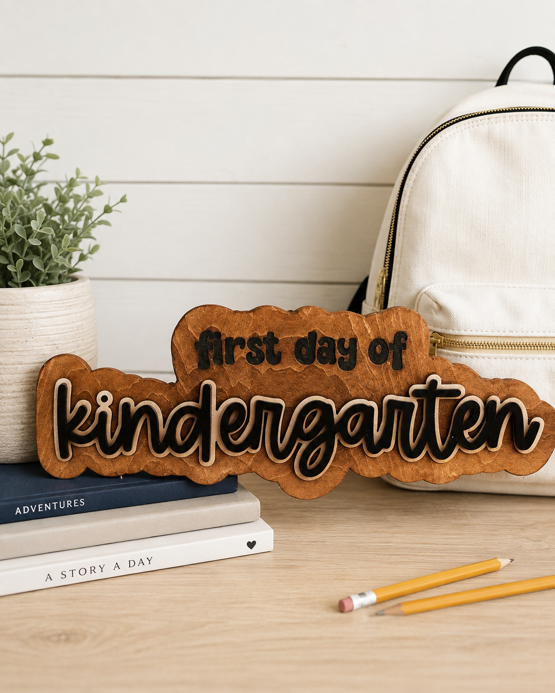 A wooden sign that reads 'first day of kindergarten' in black and white lettering, placed on a desk alongside a stack of books, a yellow pencil, a white eraser, a white backpack, and a potted green plant with leaves in a white textured pot, against a