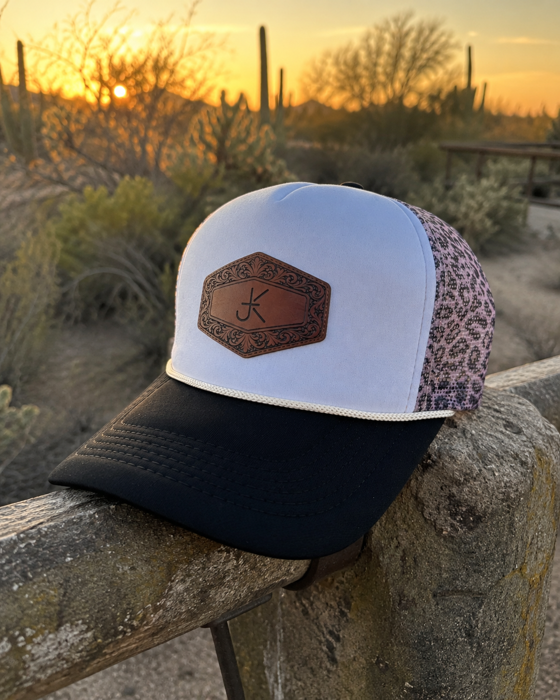 A trucker-style cap with a black brim, white front panel, and a floral patterned side panel, placed on a stone fence in a desert landscape at sunset with cacti and bushes in the background.