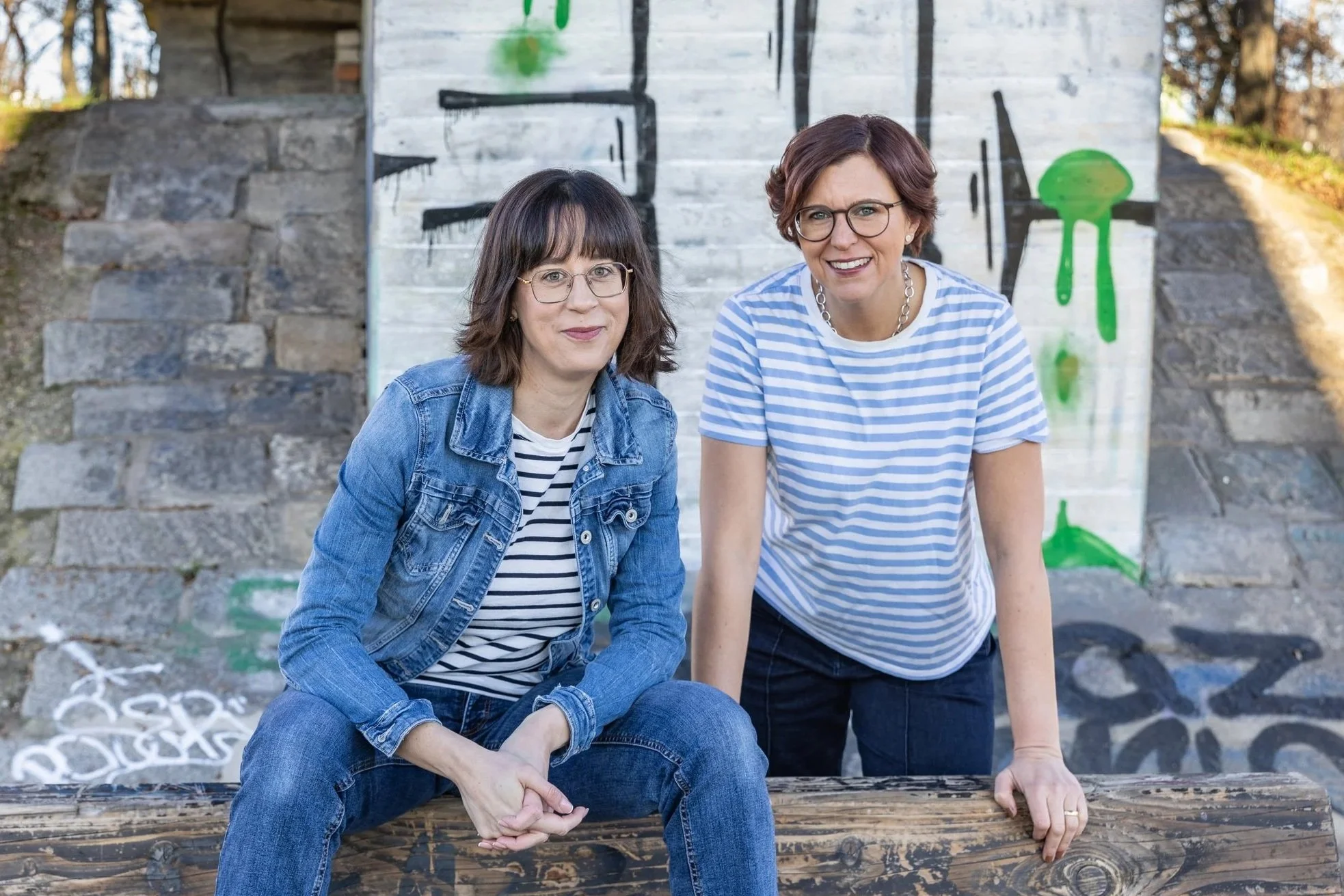 Zwei Frauen mit Brillen sitzen und stehen vor einer Wand, die mit Graffiti bemalt ist, auf einer Treppe sitzend, bei Sonnenlicht, im Freien.