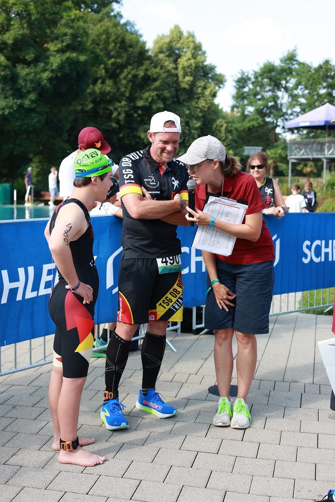 Ein männlicher Triathlet im Wettkampf w&auml;hrend eines Interviews mit einer Frau mit Mikrofon, neben einem jungen Schwimmer, im Hintergrund Badende und eine blaue Absperrung.