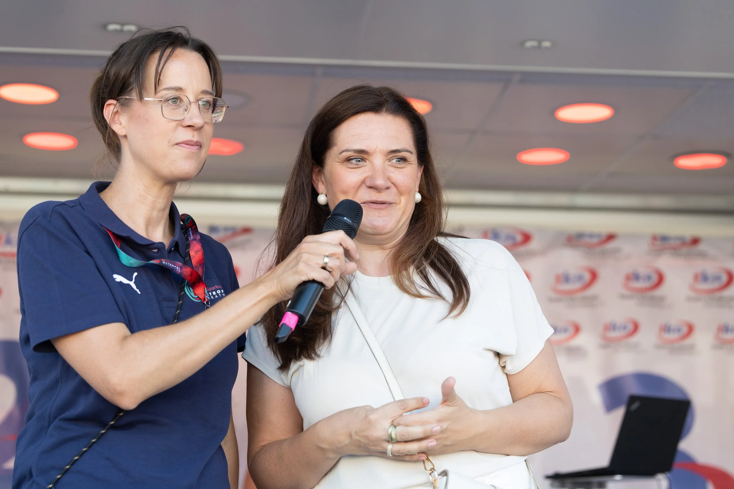 Zwei Frauen im Gespräch, eine hält ein Mikrofon, im Hintergrund ein Banner mit einem Logo.
