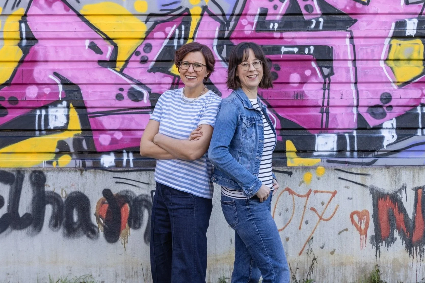 Zwei Frauen stehen vor einer bunten Graffiti-Wand, lächeln und posieren für die Kamera.