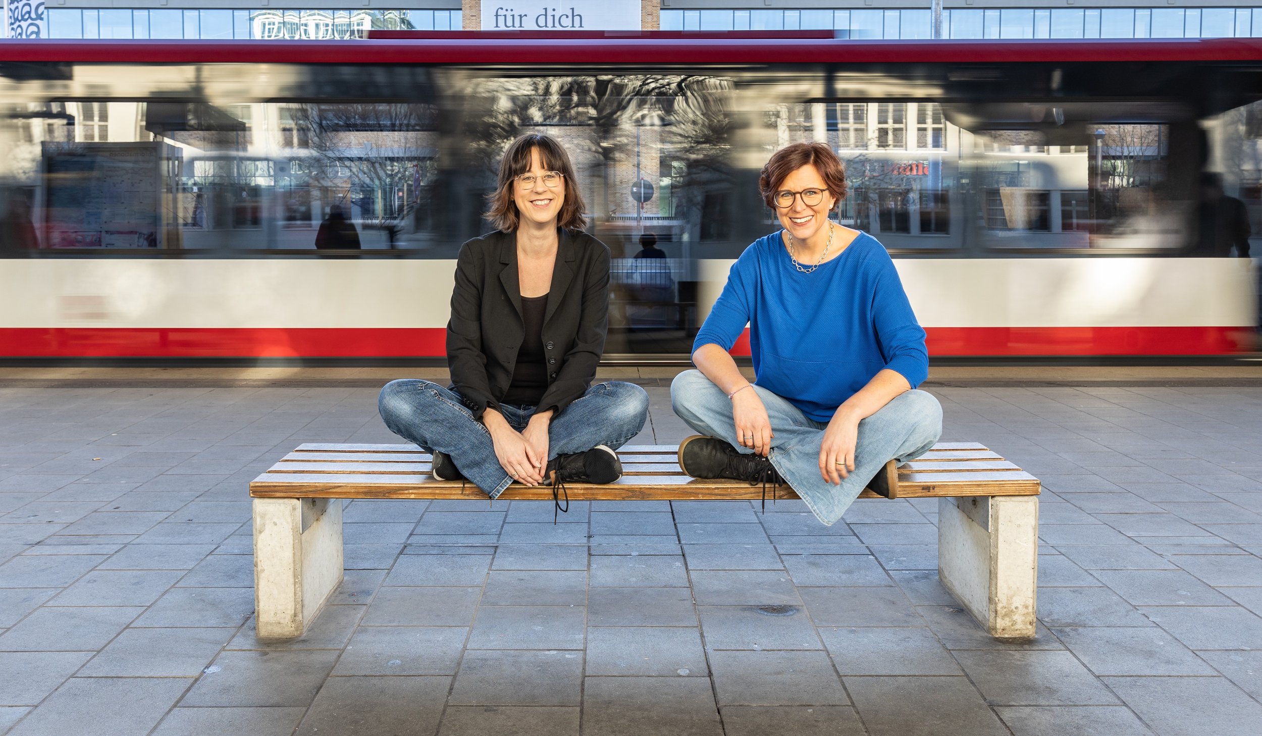 Zwei Frauen sitzen entspannt auf einer Holzbank in einer Stadt, im Hintergrund fährt ein Bus vorbei.