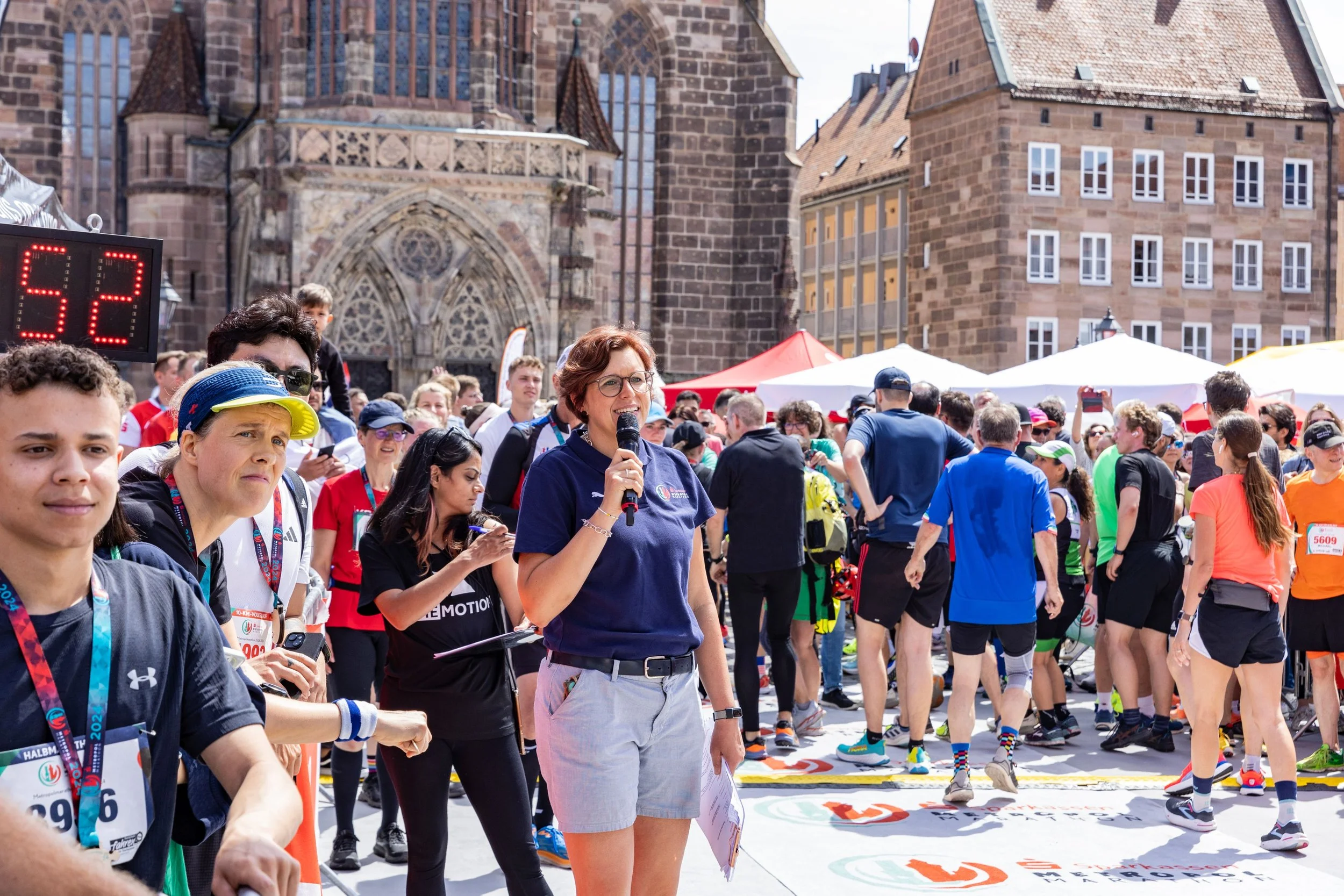 Menschen bei einem Marathon oder Straßenlauf, einige halten Medaillen, eine Person spricht in ein Mikrofon, im Hintergrund sind historische Gebäude und Zelte, es ist sonnig.