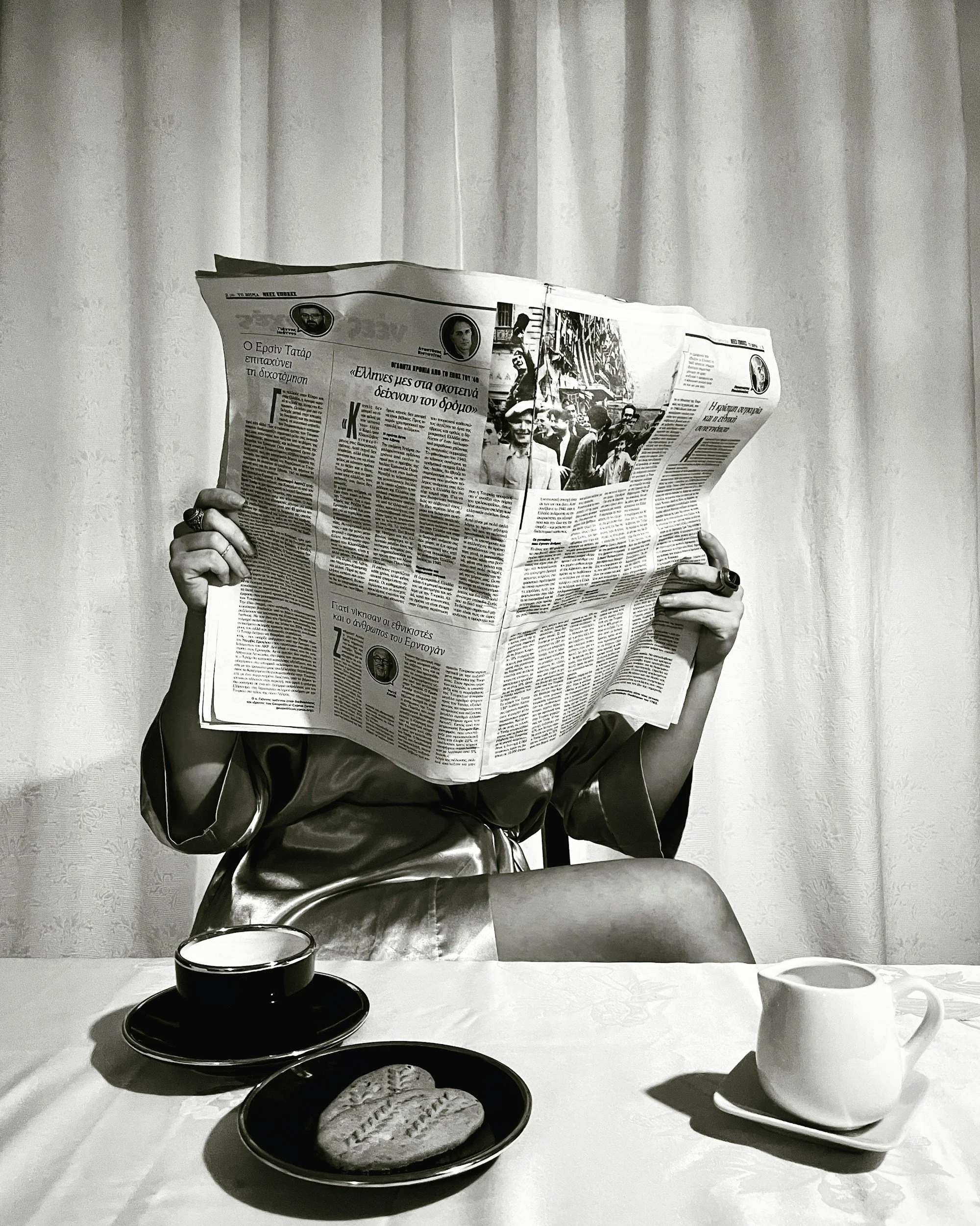 A person is sitting at a table with a tablecloth, holding an open newspaper that covers their face. On the table, there are two white cups, one of which is on a saucer. In front of the person, there are two black plates, one with a cookie or biscuit. The background features plain curtains.
