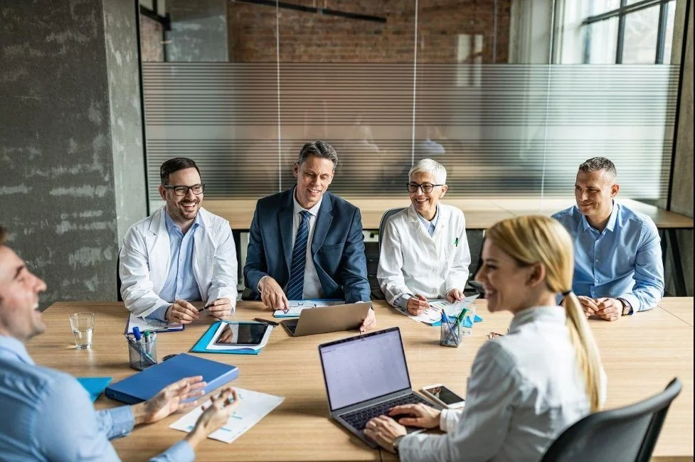 Business meeting with six professionals seated around a conference table, engaged in discussion and smiling, in a modern office setting.