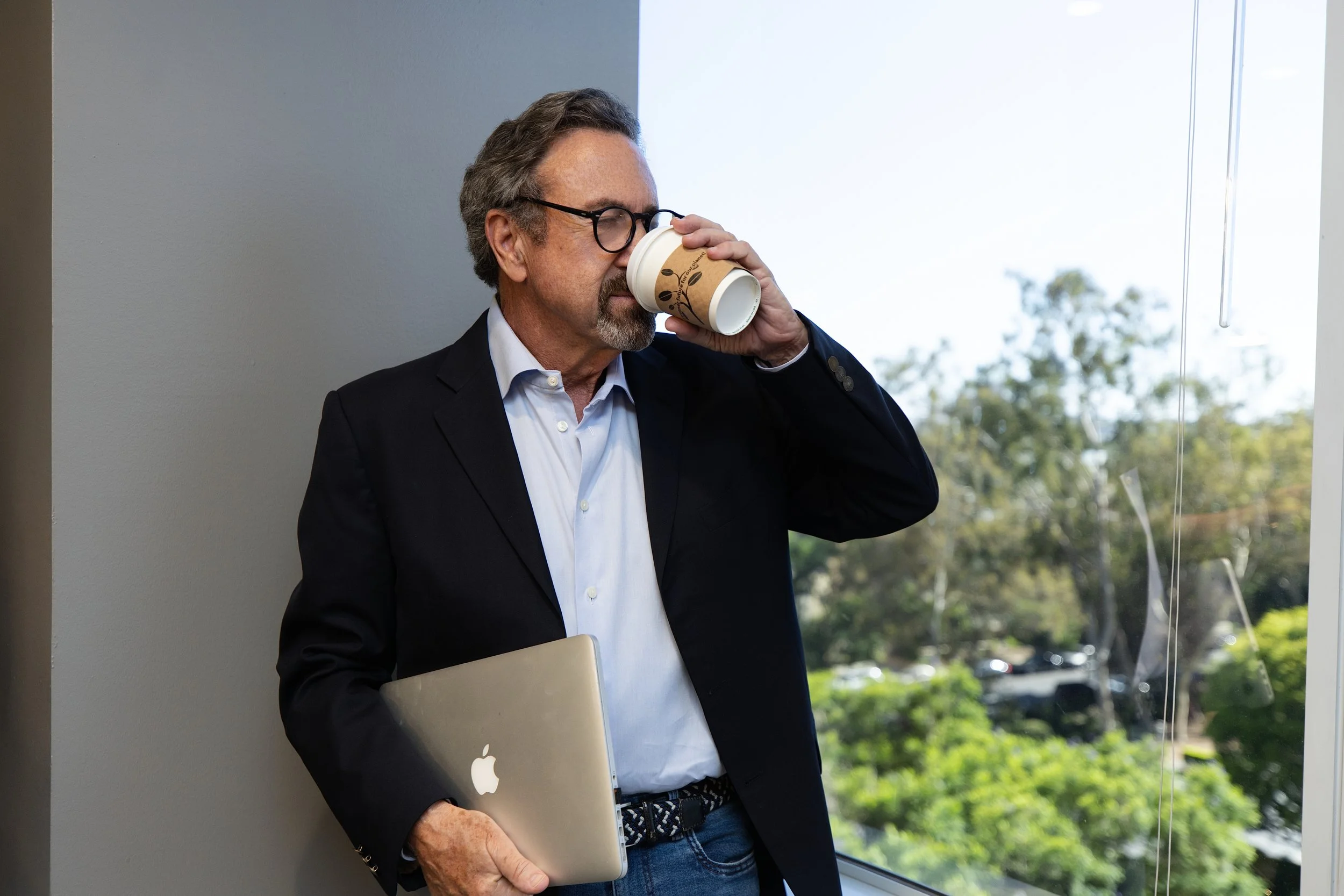 A middle-aged man with gray hair, beard, and glasses wearing a dark blazer and white shirt, holding a laptop and drinking coffee from a disposable cup, standing near a window with outdoor trees visible.