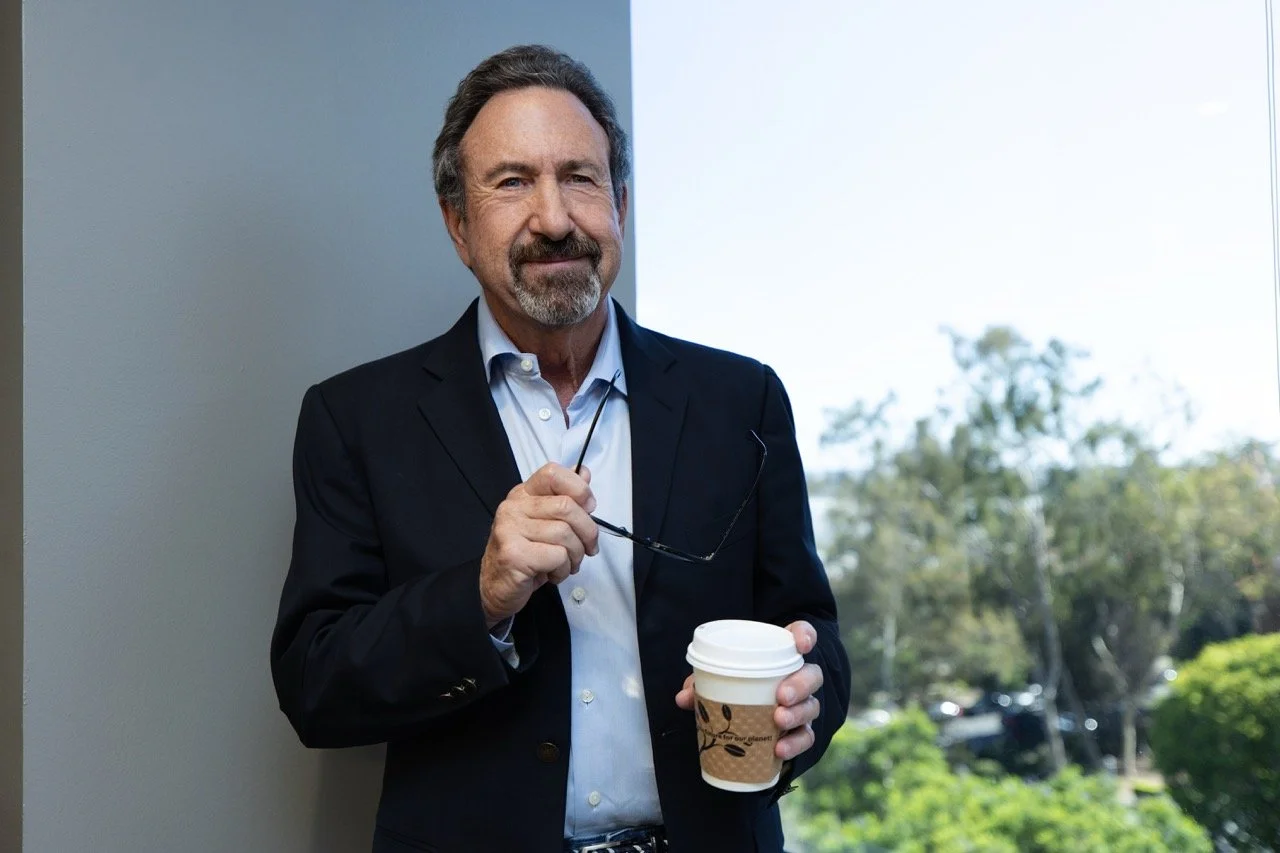 A middle-aged man with gray hair and a beard, wearing a dark suit jacket and a light blue shirt, standing indoors near a window. He is holding a disposable coffee cup in one hand and a pair of glasses in the other. Outside the window, trees and a parking lot are visible.