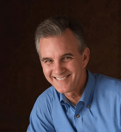 Man with short gray hair in a blue button-up shirt smiling against a dark brown background.