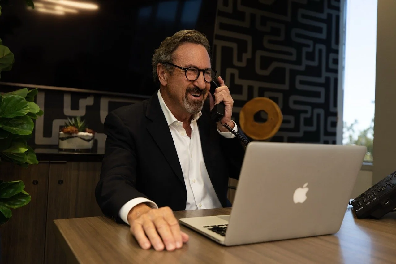 A middle-aged man with glasses and gray hair, wearing a black suit and white shirt, sitting at a wooden desk in an office. He is smiling and talking on a corded landline phone while looking at his open MacBook laptop. There is a potted plant on the left and a decorative object on the right of the man, with a window in the background.