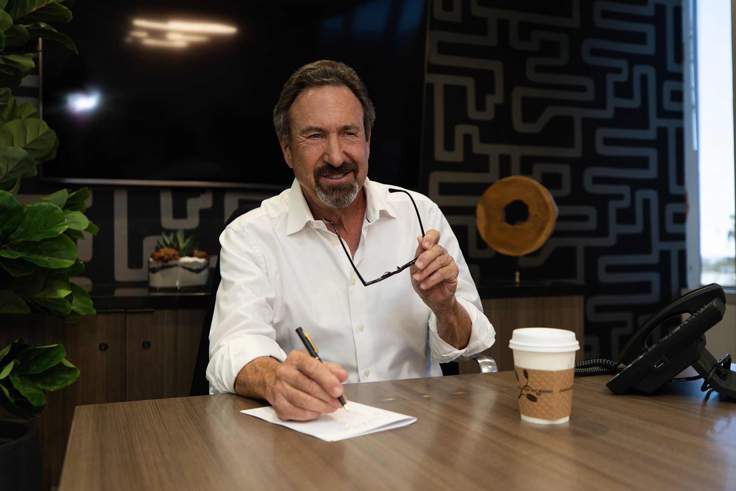 A middle-aged man with gray hair and a beard, wearing a white shirt, sitting at a conference table with a cup of coffee, paper, and pen, smiling and holding eyeglasses, in a modern office with dark walls, plants, and a large television screen.