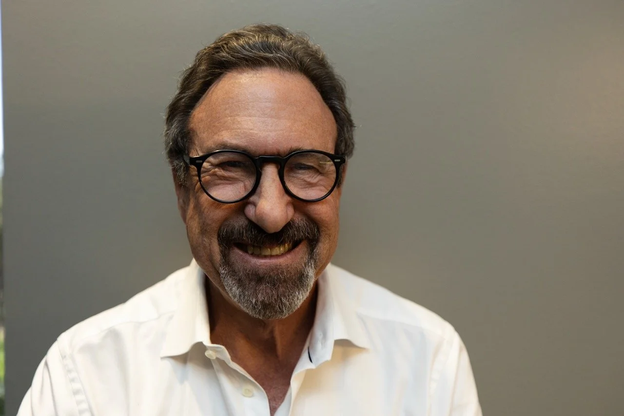 A smiling middle-aged man with dark hair, glasses, a beard, and a mustache, wearing a white collared shirt, standing against a plain gray background.