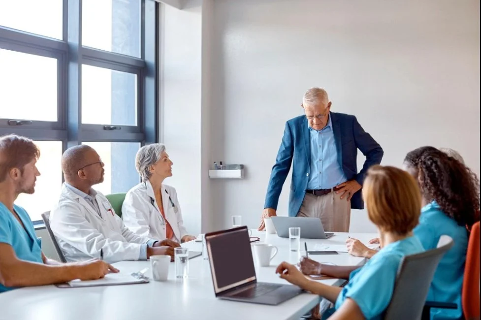 A meeting room with seven medical professionals, including doctors and nurses, listening to an older male speaker standing at the head of the table with a laptop. The room has large windows and natural lighting.