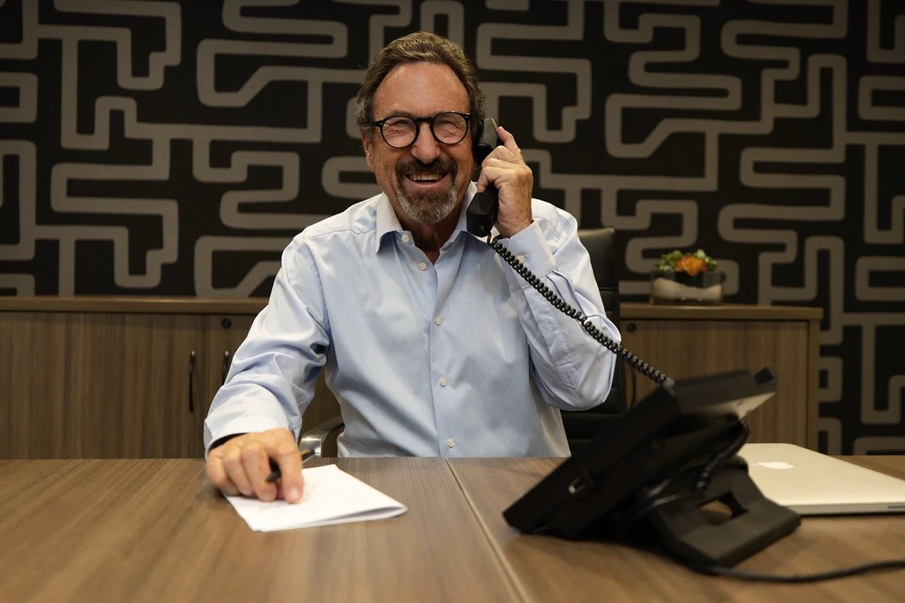 A man with glasses, gray hair, and a beard, wearing a white dress shirt, is sitting at a desk in an office, talking on a black landline phone while smiling and laughing.