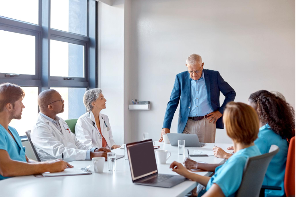 A meeting room with seven medical professionals, including doctors and nurses, listening to an older male speaker standing at the head of the table with a laptop. The room has large windows and natural lighting.