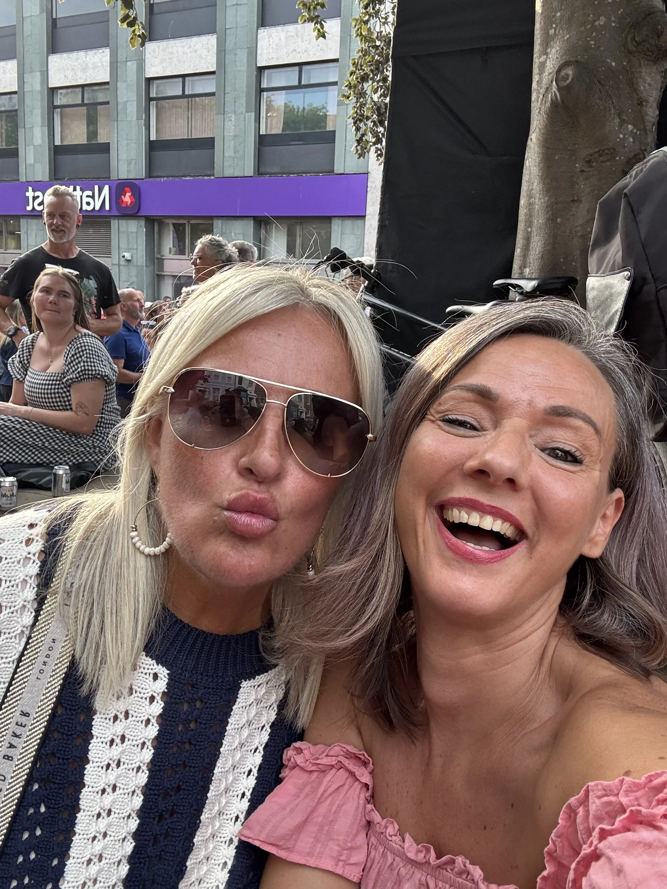 Close-up of two smiling women taking a selfie at an outdoor event with a crowd and buildings in the background.
