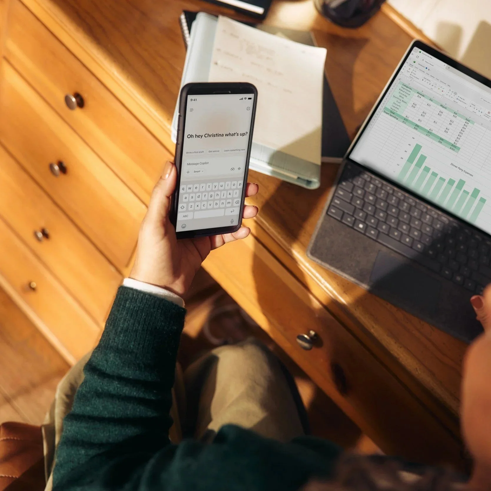 Person holding a smartphone displaying a message that says 'Oh hey Christina, what’s up?' while sitting at a wooden desk with a laptop showing a spreadsheet and a notebook.