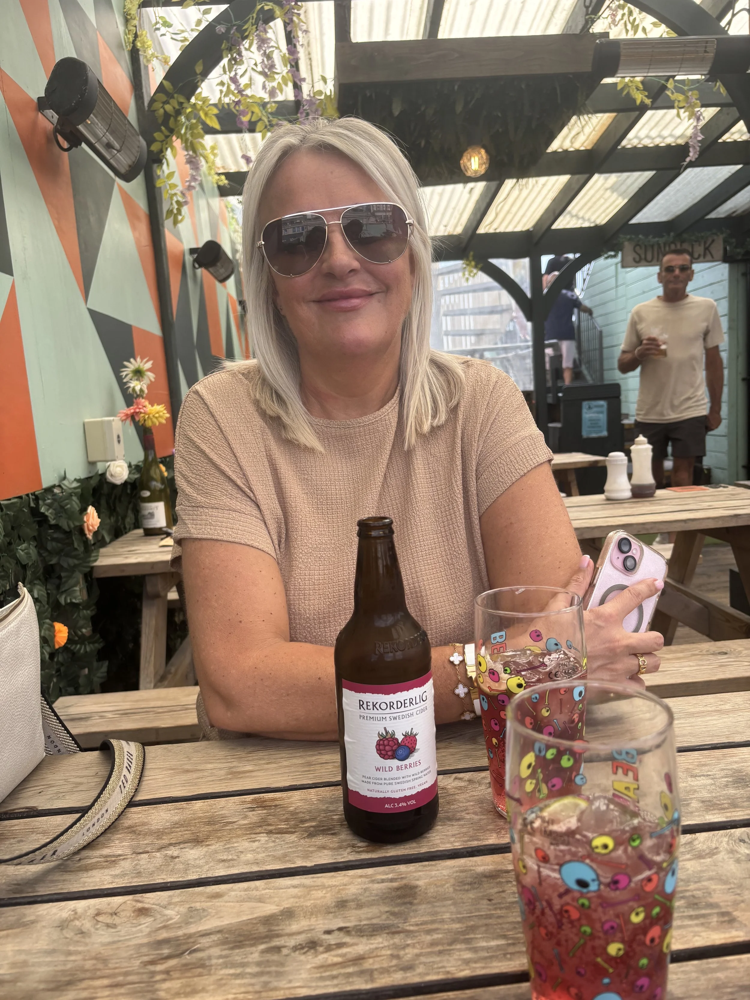 A woman with blonde hair wearing sunglasses and a beige shirt, smiling, sitting at a wooden table at an outdoor cafe. She is holding a phone in her right hand. On the table are a brown bottle of Rekorderlig Wild Berries cider, a glass with colorful emoji-themed decorations, and other glasses. In the background, there's a man in a white t-shirt and sunglasses holding a drink, standing near a green building, with a shaded pergola and some string lights overhead.