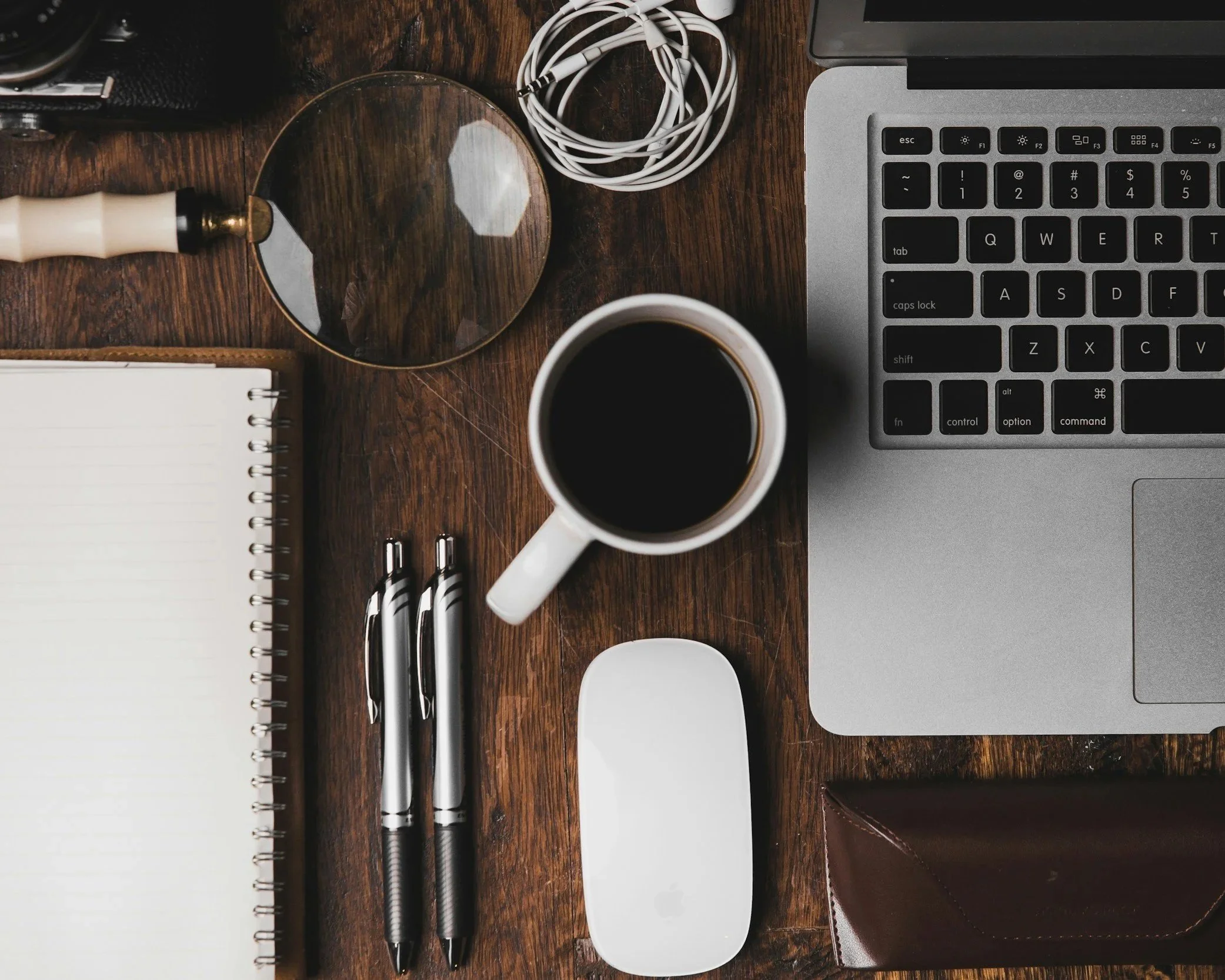 A top-down view of a wooden desk with various objects including a camera, a mirror, earbuds, a cup of coffee, pens, a notepad, and a laptop keyboard.