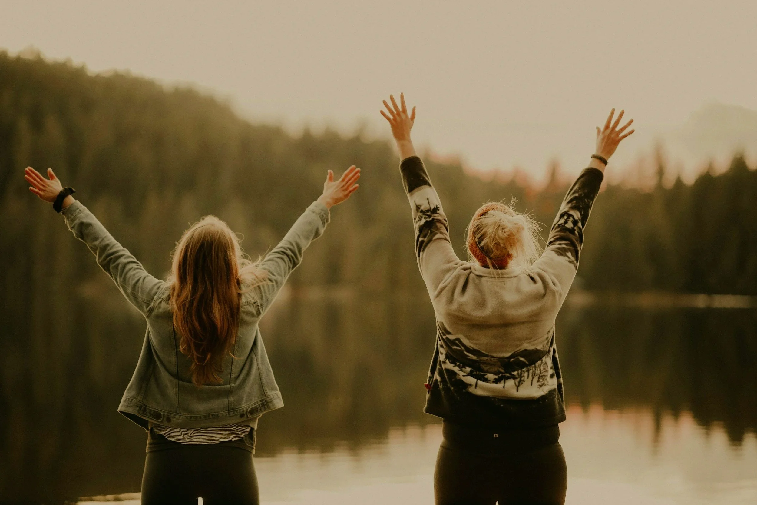 Two women with arms raised standing near a lake, with a blurred forest in the background.