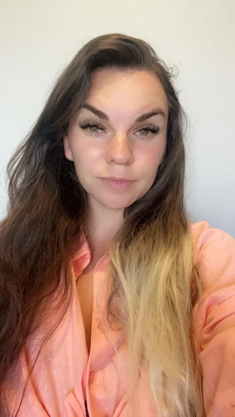 A young woman with long, wavy brown hair and light-colored eyes, wearing a light pink shirt, standing against a plain light-colored background.