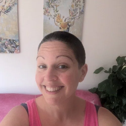 A woman with short hair smiling and wearing a pink tank top, sitting on a pink couch, with artwork and a green plant in the background.