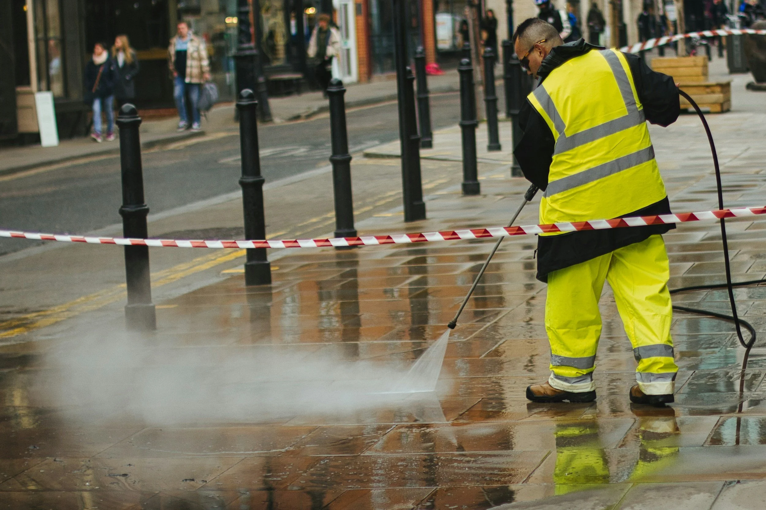 A person wearing a yellow safety vest and yellow pants is pressure washing a sidewalk. There are black posts along the street and people in the background walking along the sidewalk.