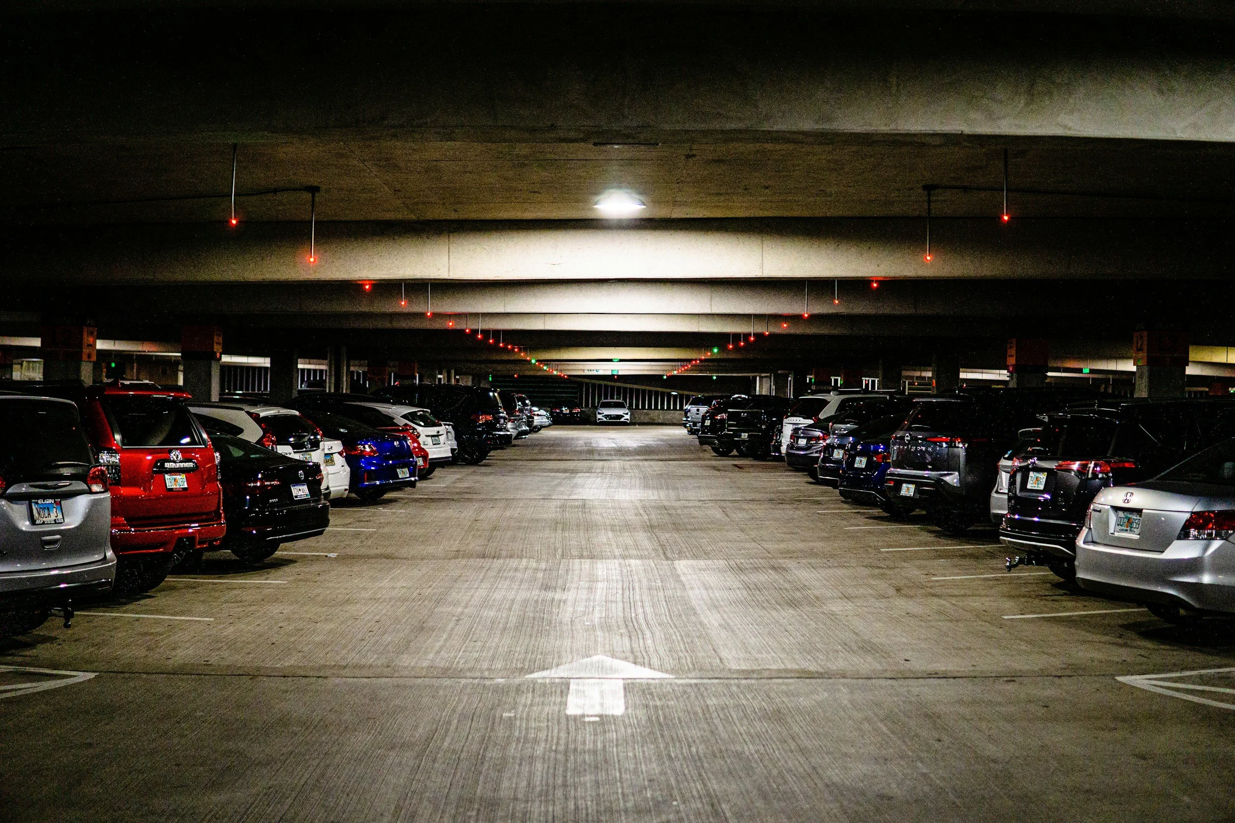 Underground parking garage with rows of parked cars beneath a concrete ceiling. Red and green lights are installed on the ceiling, and a white arrow painted on the floor points forward.