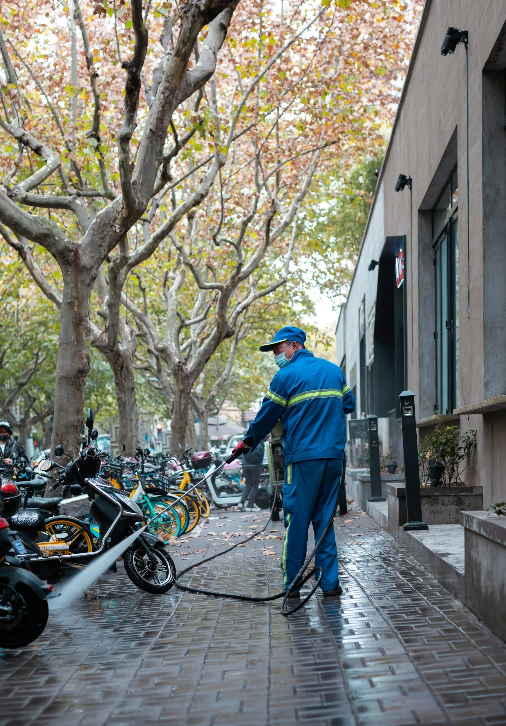 A worker in blue uniform and face mask is cleaning a sidewalk with a high-pressure hose, with parked motorcycles and bicycles lining the street and trees with fall foliage in the background.
