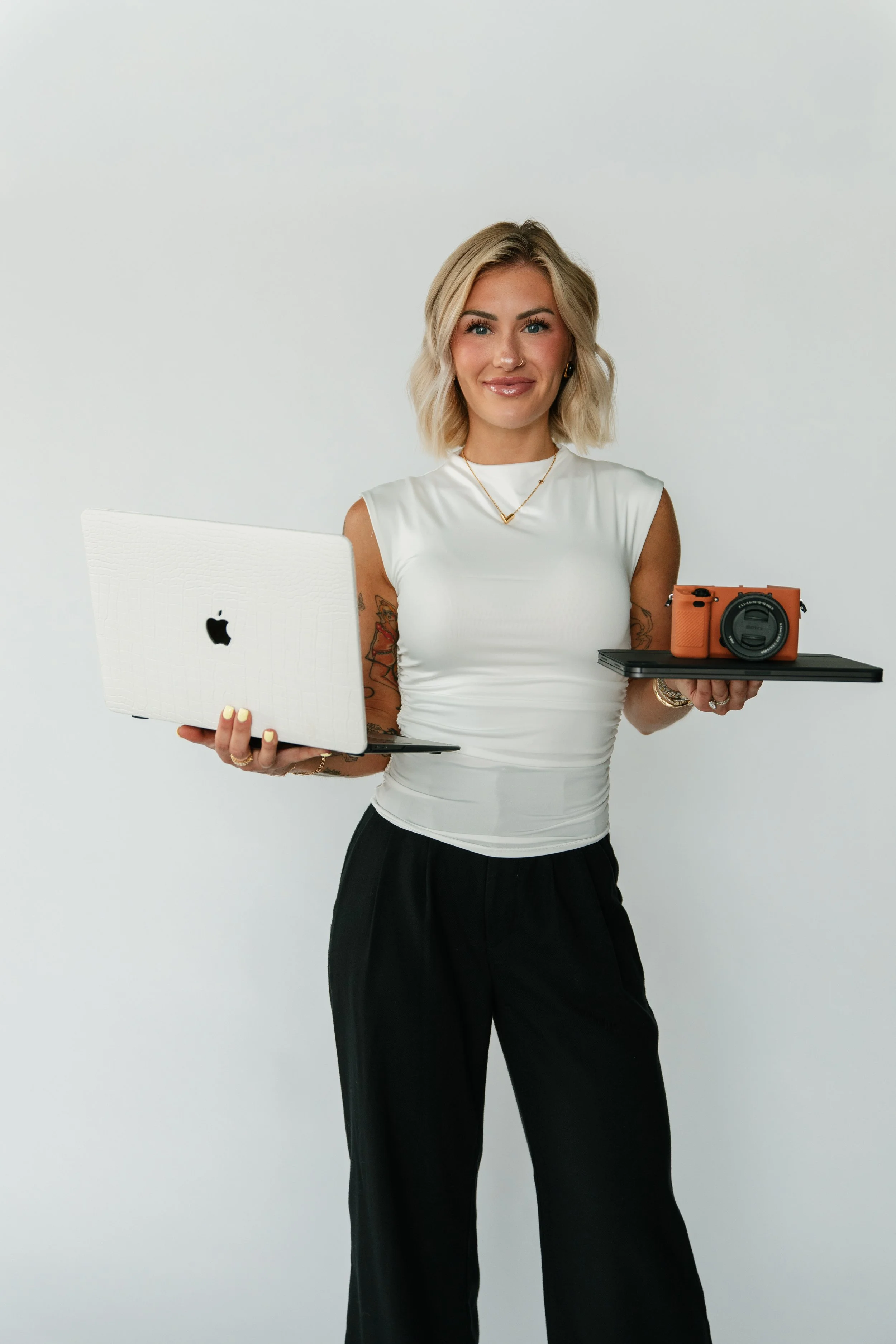 A woman with blonde hair holding a laptop in one hand and a tray with a camera on it in the other, standing against a plain white wall.