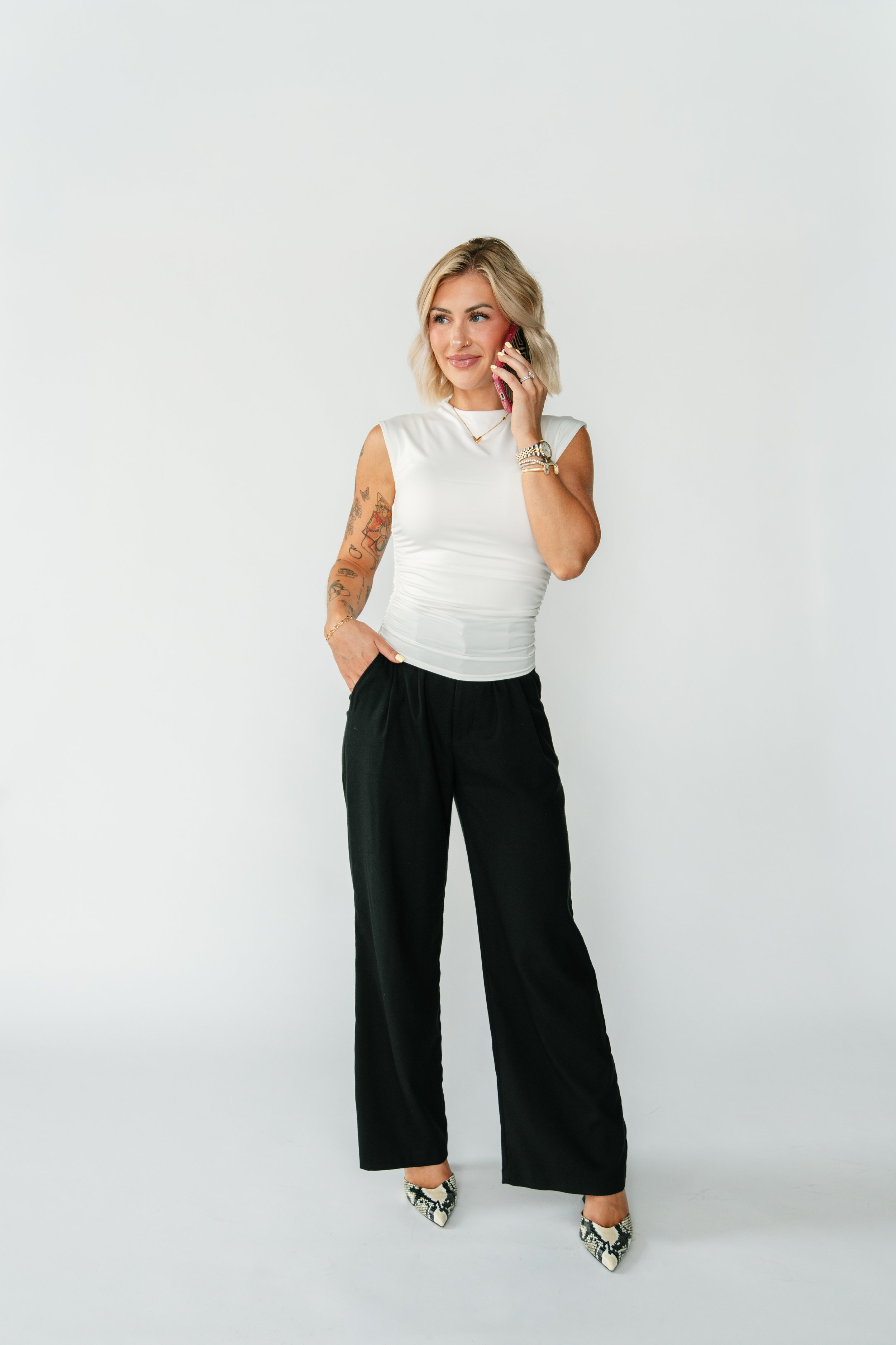 A woman with blonde hair wearing a white sleeveless top, black pants, and high heels, standing against a plain white background, talking on her cellphone.