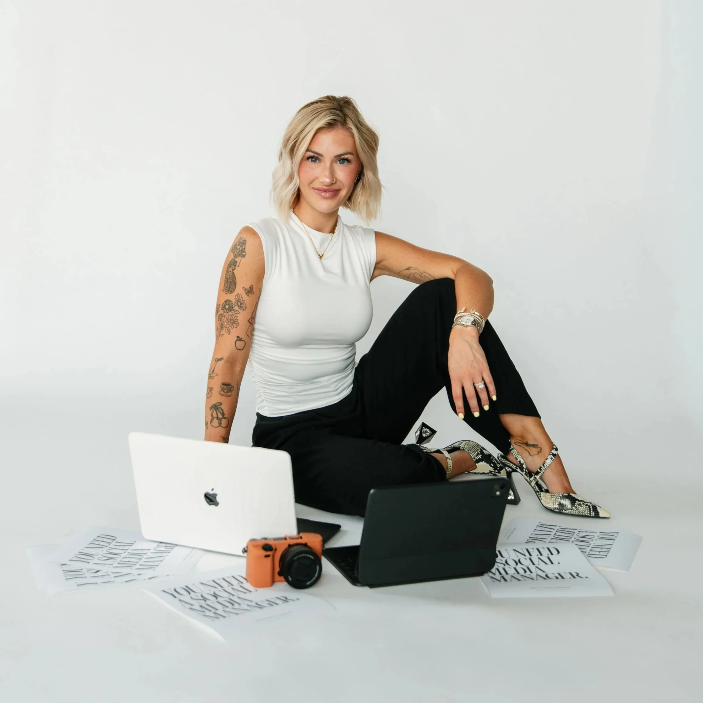 Woman sitting  surrounded by papers, a camera, and a laptop in a minimalist room with a plain white wall background.