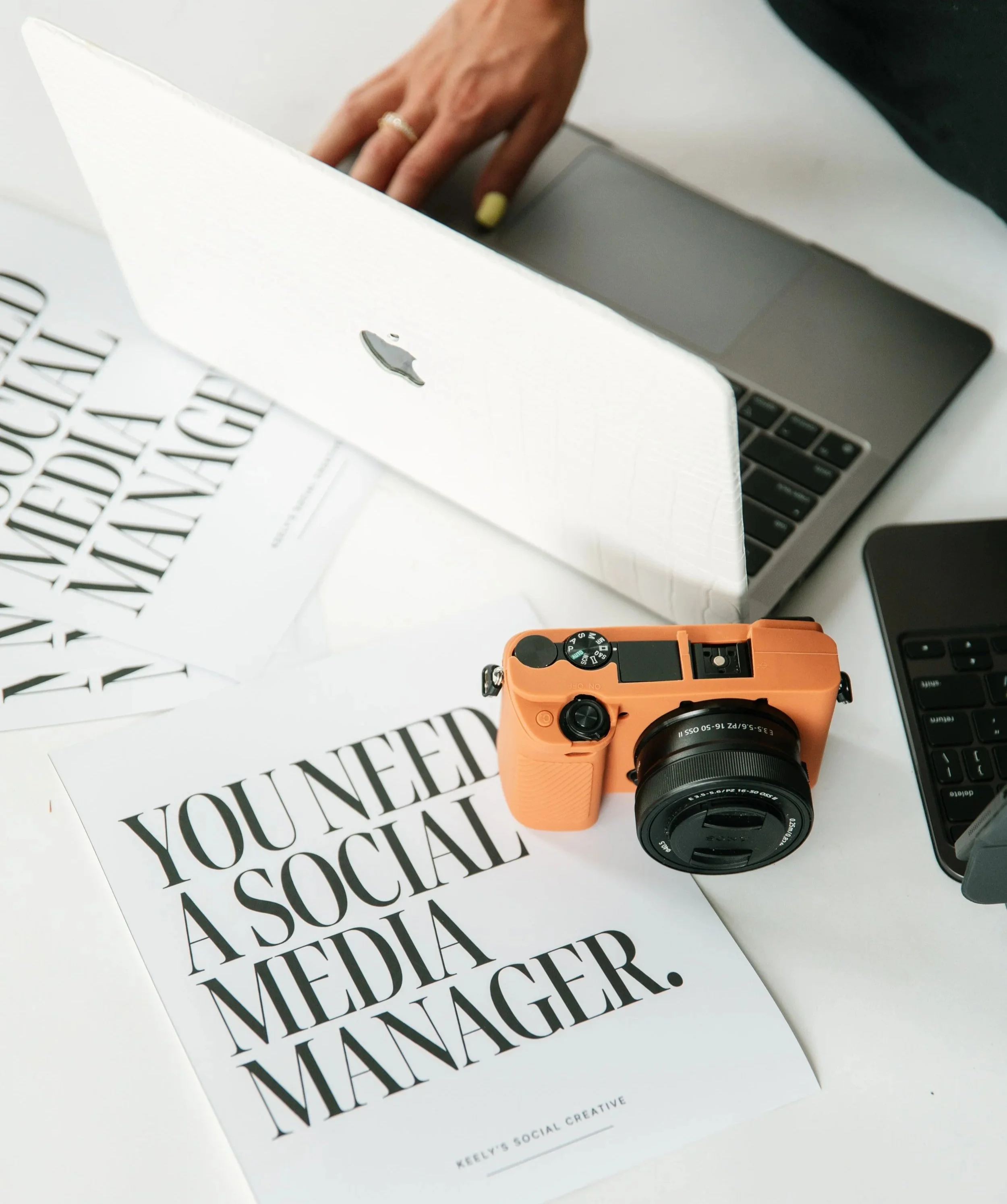 Close-up of a person's hand resting on a laptop keyboard with a white paper that has bold, large text reading 'YOU NEED A SOCIAL MEDIA MANAGER,' and an orange camera placed nearby.