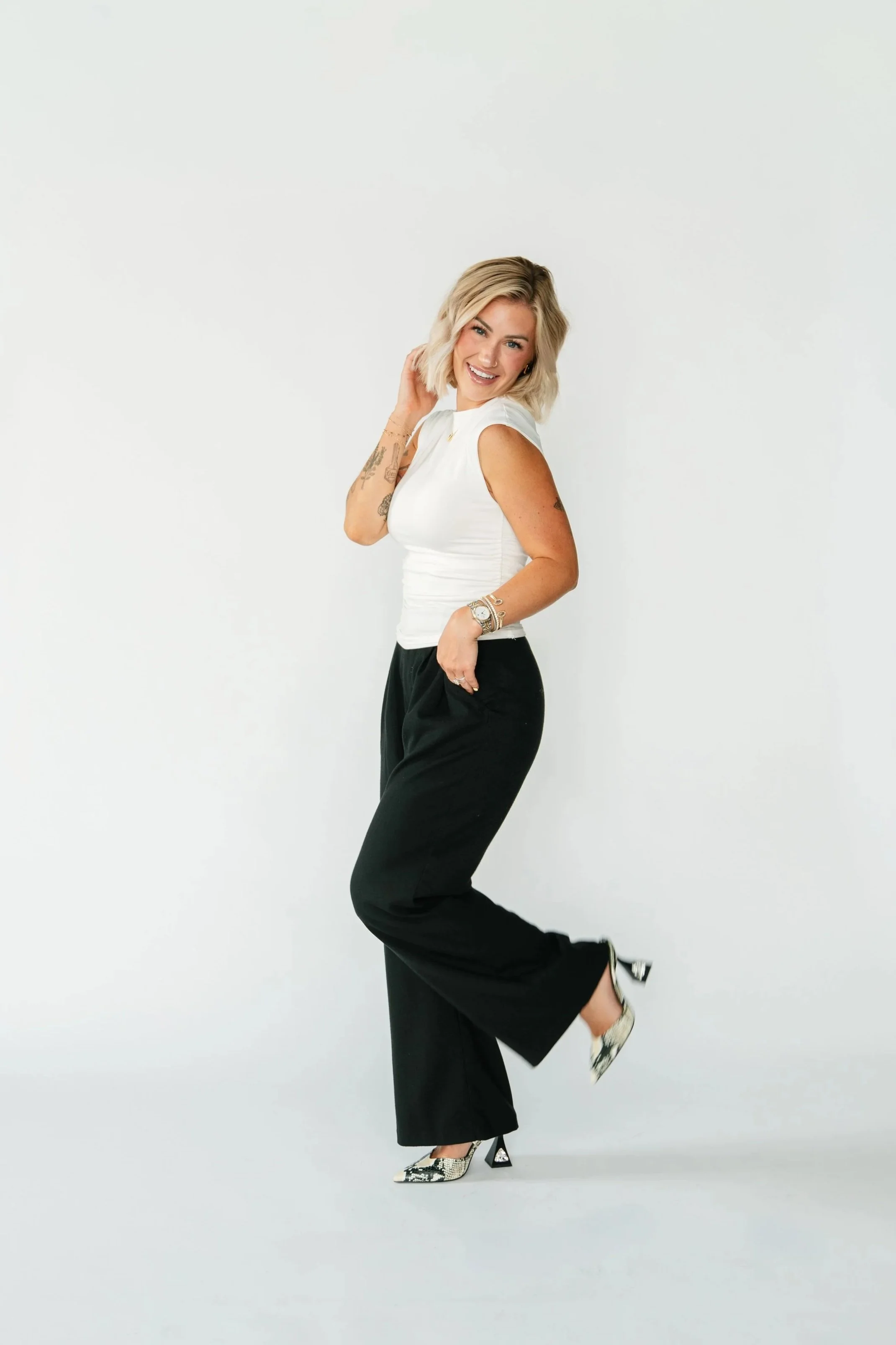 A woman with blonde hair is smiling and posing on one leg, wearing a white sleeveless top, black wide-leg pants, and high heels against a plain white background.