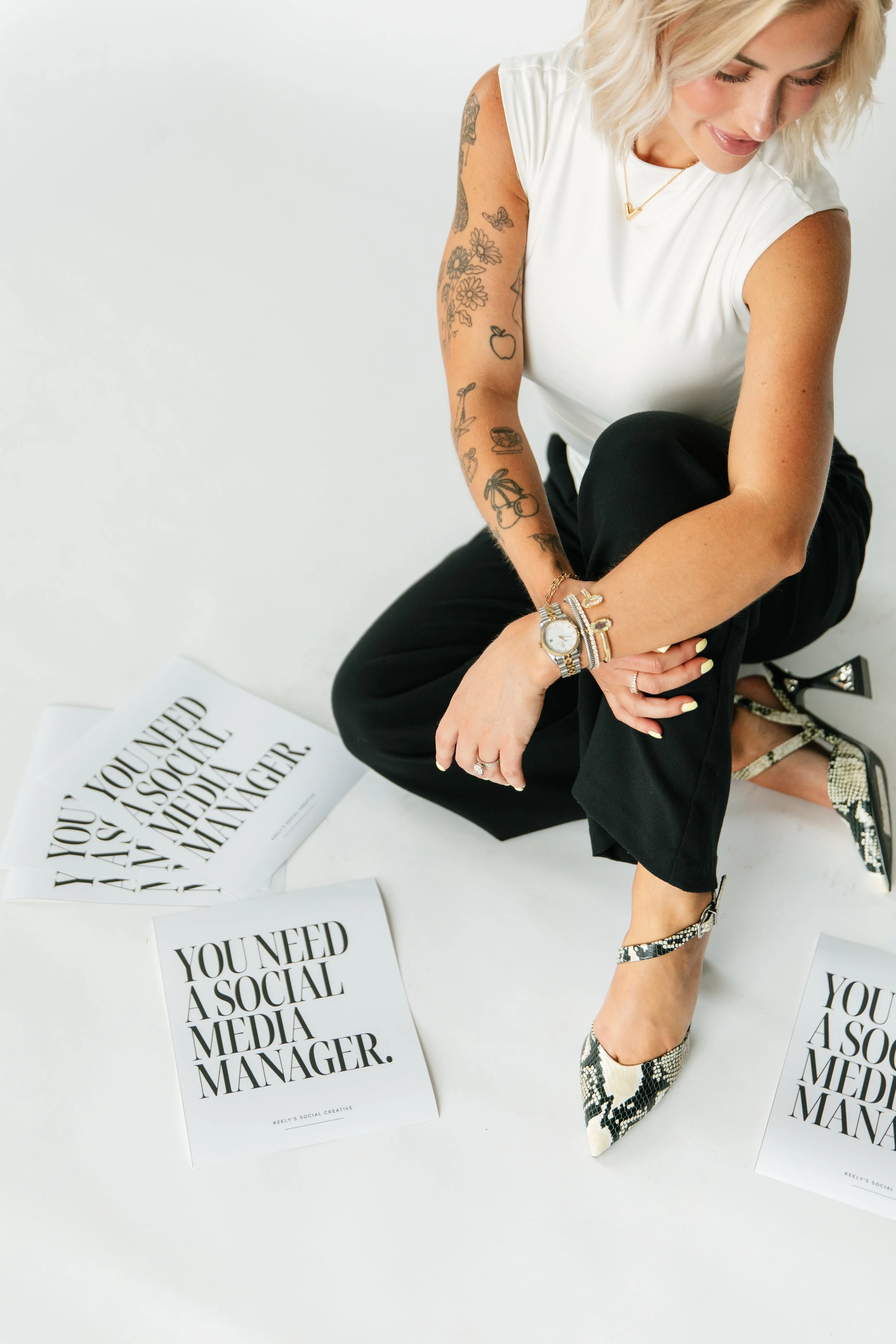 A woman with blonde hair seated on the floor surrounded by printed papers that read "You need a social media manager." She is wearing a white sleeveless top and black pants.