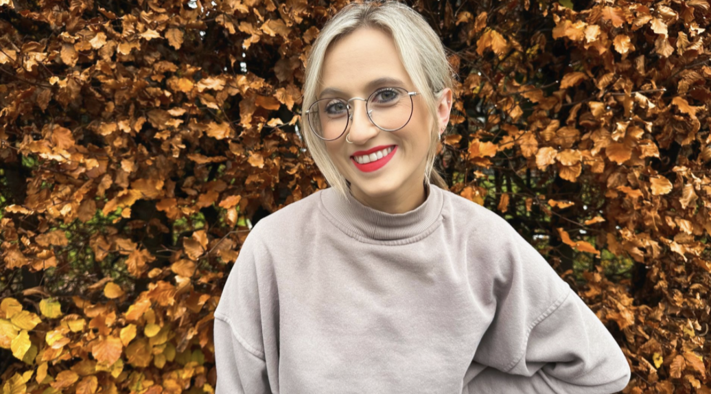 A smiling woman with blonde hair and glasses standing outdoors in front of an autumn-colored bush.