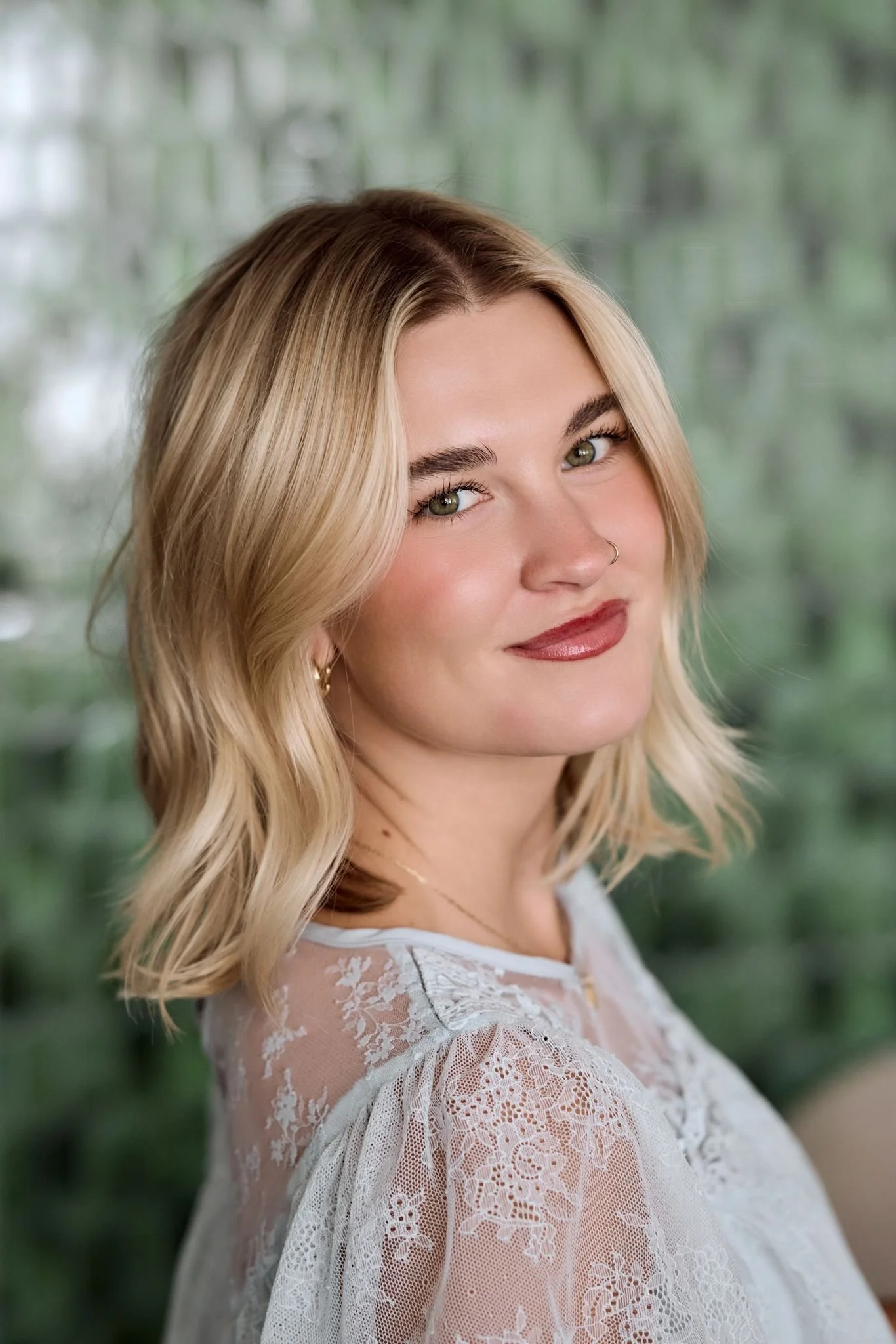 Close-up of a woman with blonde wavy hair, green eyes, fair skin, wearing a nose ring, gold earrings, and a white lace top, standing outdoors with a blurred green background.