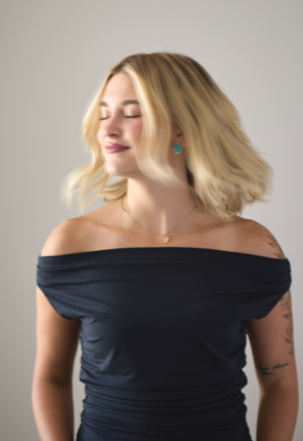 A smiling woman with blonde hair, wearing a black off-the-shoulder top, turquoise earrings, and a gold necklace, standing against a plain background.