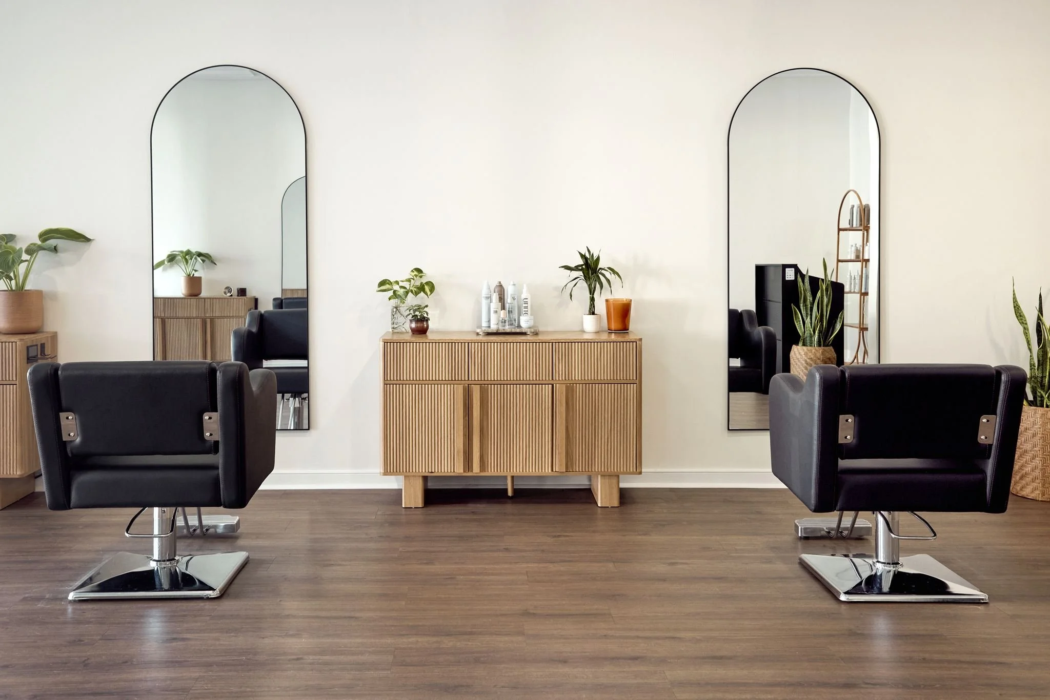 Modern hair salon with two black styling chairs, a wooden cabinet with potted plants and hair products, large mirrors, and decorative plants on the wall.