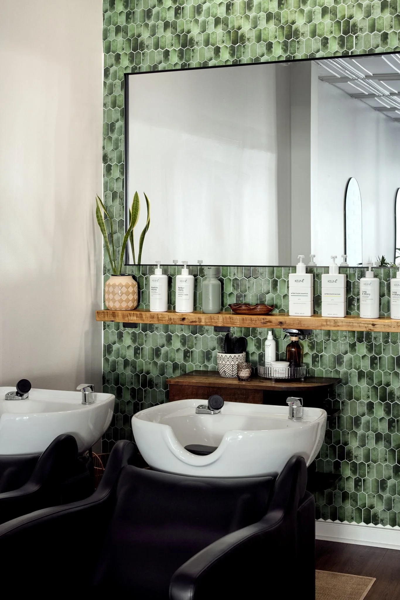 A bathroom with two white sinks, a large mirror, and a wooden shelf holding bottles of hair and skincare products. The wall behind the mirror is decorated with green hexagonal tiles, and a potted plant with long, pointed leaves sits on the left side of the shelf.