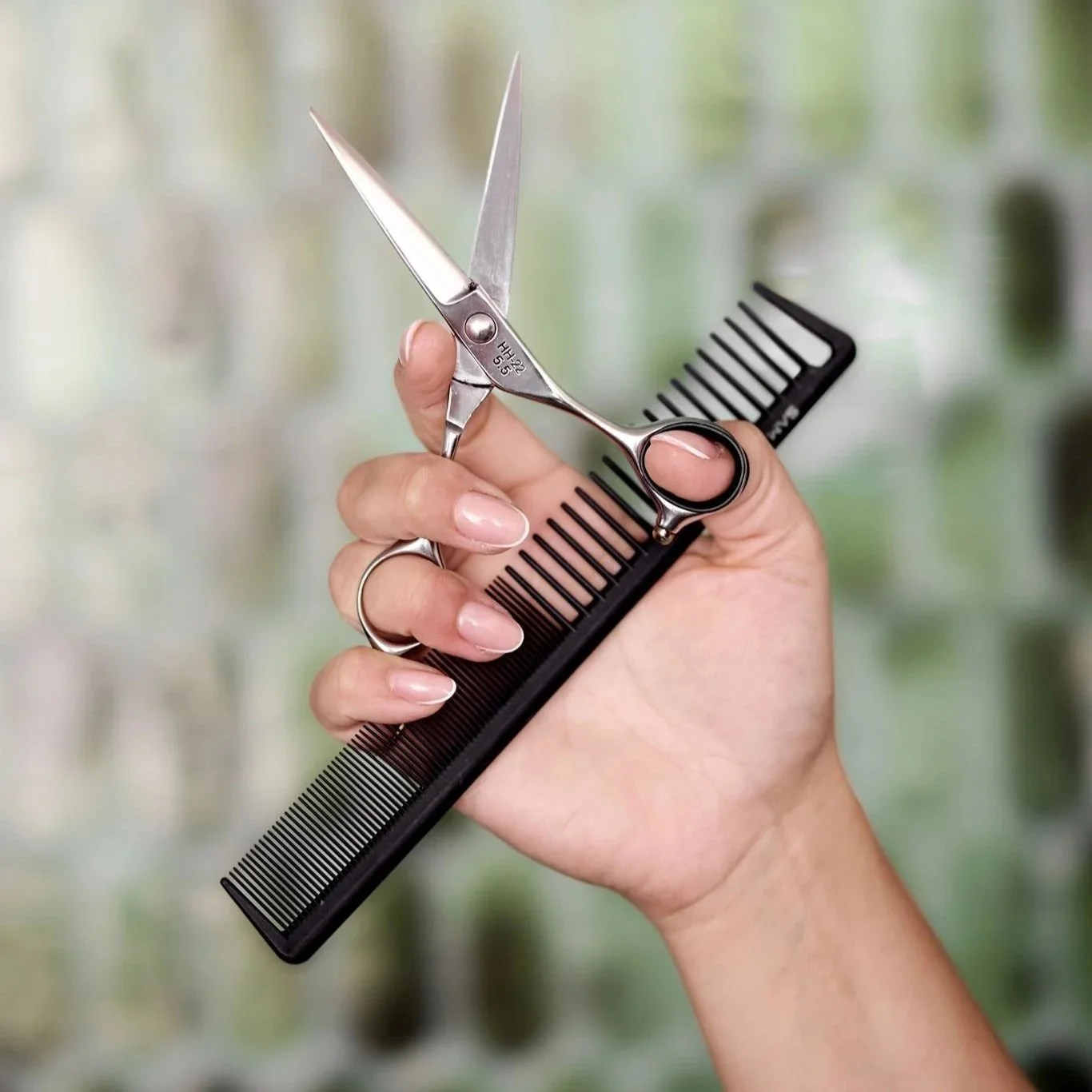 A person's hand holding a pair of scissors and a black hair comb, with a blurred green and white background.