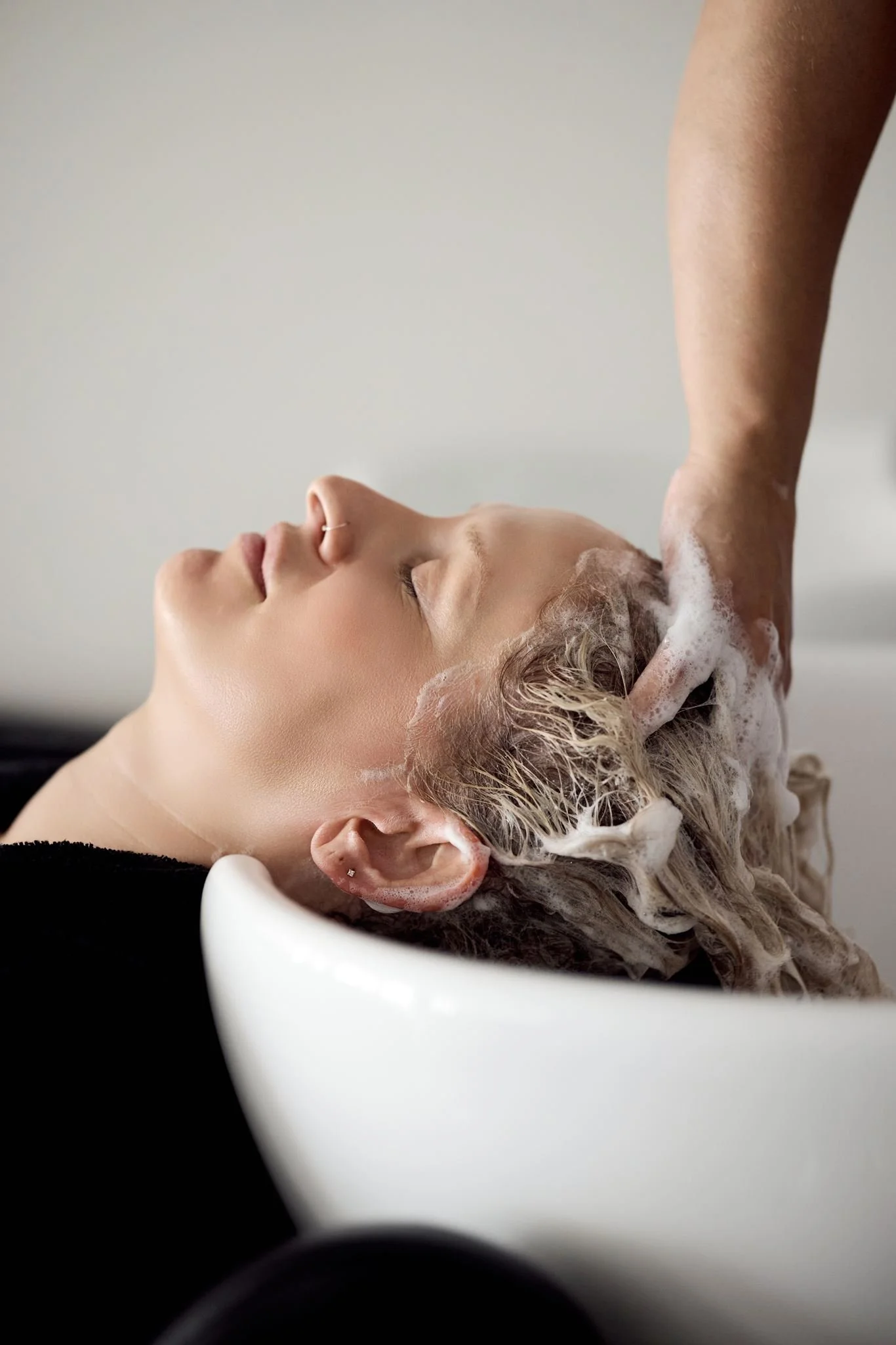 A woman reclining with her eyes closed, receiving a head massage or hair wash in a salon, with soap bubbles and foam on her hair and a white basin supporting her head.