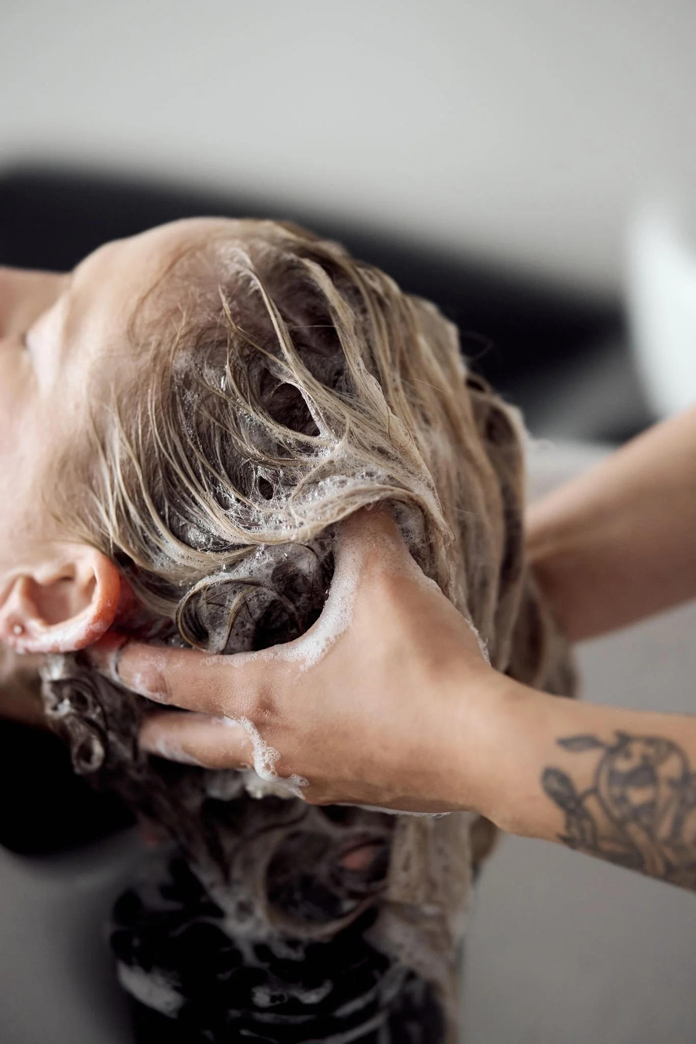 Close-up of a person with blonde, curly hair being shampooed, with soap lather and a tattooed arm visible.