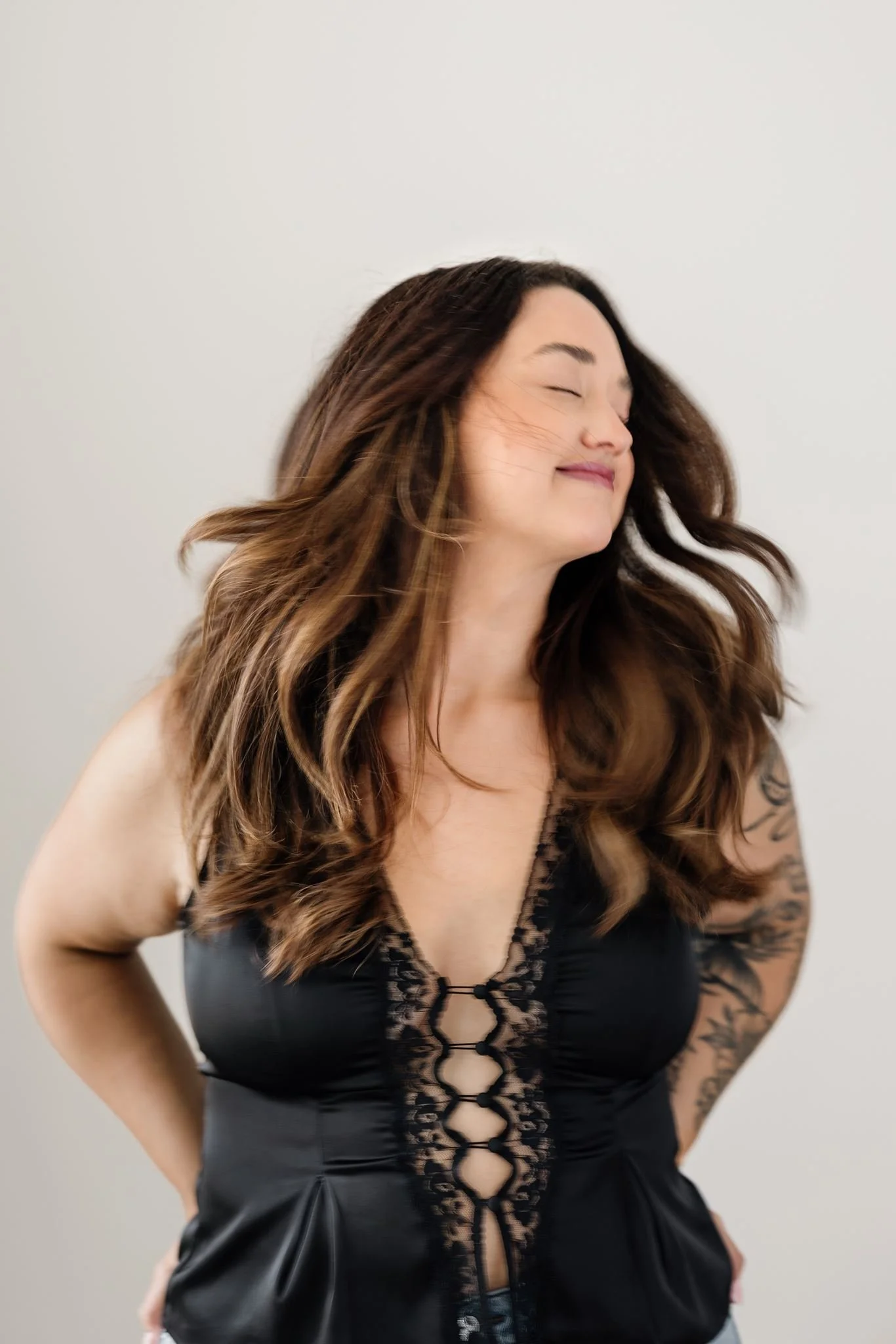 A woman with long wavy brown hair, smiling with eyes closed, wearing a black lace-up top with lace details and a tattoo on her left arm, posing against a plain light-colored background.