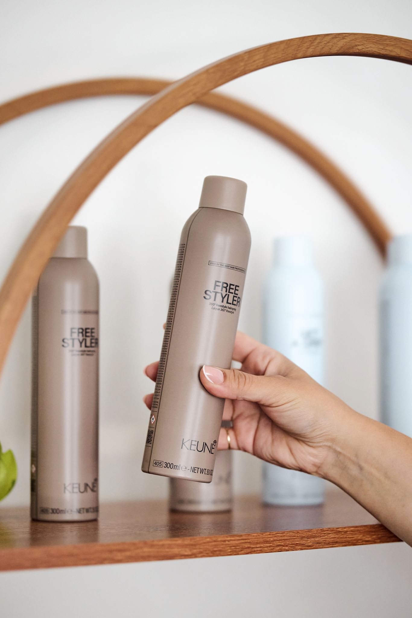 Hand holding a beige bottle of KEUNE Free Style hair product, with three more bottles on a wooden shelf behind. The bottles are similar in color and design.